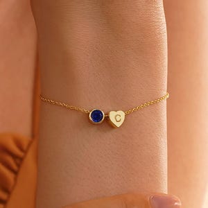 Könnte beinhalten: Ein zartes Goldkettenarmband mit einem kleinen, runden, dunkelblauen Edelsteinanhänger und einem winzigen herzförmigen Goldanhänger, der mit dem Buchstaben C graviert ist. Das Armband besteht aus einer dünnen, polierten goldfarbenen Kette.
