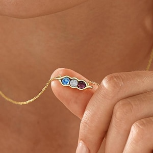 May include: A gold chain necklace featuring a horizontal pendant shaped like a pea pod containing three round gemstones in blue, clear, and dark pink. The delicate metal chain connects to each side of the small, curved, pod-like setting.
