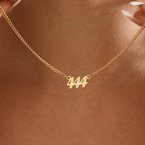 Könnte beinhalten: Eine goldfarbene Halskette mit einem Anhänger mit der Zahl "444". Die Halskette wird von einer Person hochgehalten. Die Kette ist filigran und der Anhänger ist in einer Schriftart geschrieben. Der Hintergrund ist unscharf.
