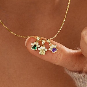 May include: A gold chain necklace featuring three paw print pendants. Each pendant is accented with small clear stones and a central heart-shaped gemstone in green, yellow, and purple. The necklace is displayed on a delicate gold link chain.