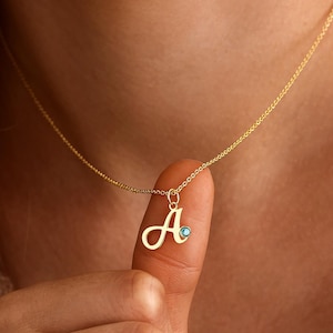 Pode incluir: Um colar de tom dourado com uma corrente fina e um pingente com a letra A em cursivo. O pingente é adornado com uma pequena pedra preciosa redonda de cor azul claro incrustada no metal. O texto LeslieArtJewelCo é visível no canto inferior direito.