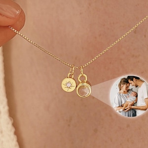 Könnte beinhalten: Eine goldfarbene Halskette mit zwei kleinen Anhängern. Ein Anhänger ist ein runder Charm mit einem Sternmuster und einem kleinen Edelstein. Der andere Anhänger ist ein klarer, runder Charm. Im Hintergrund ist ein kleines Familienbild.
