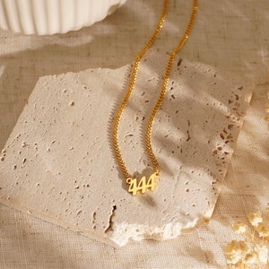 May include: A gold-colored necklace with the number 444 pendant. The necklace is displayed on a textured, light-colored stone surface. The chain is delicate and the pendant is in a serif font.