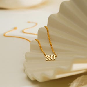 May include: A gold-colored necklace with a delicate chain and a pendant featuring the number "333". The necklace is displayed on a white, textured surface. The jewelry is simple and elegant.