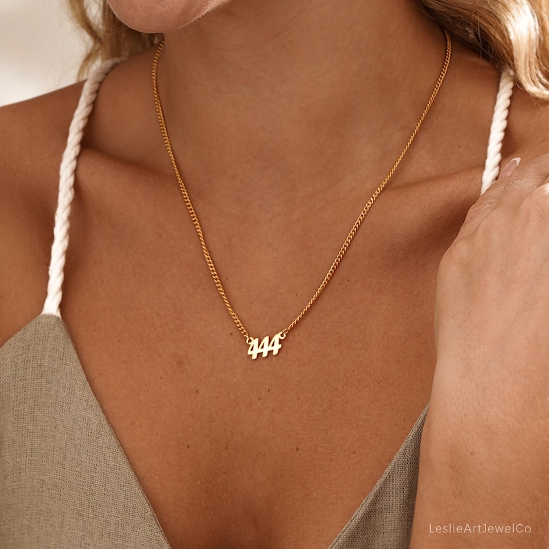 May include: A gold-colored necklace with a delicate chain and the number "444" as a pendant. The necklace is worn around the neck. The image is a close-up shot.