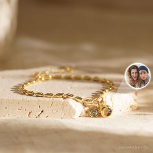 Könnte beinhalten: Ein goldenes Kettenarmband mit kleinen ovalen Gliedern und zwei Anhängern. Ein Anhänger ist ein kleiner runder, klarer Kristall, der andere ist ein größerer runder Anhänger mit einem klaren Kristall im Inneren. Das Armband liegt auf einer hellbraunen Oberfläche.
