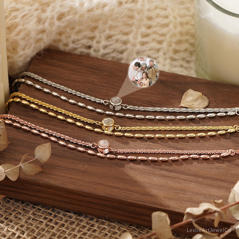 May include: Three layered bracelets in silver, gold, and rose gold tones, each featuring a small circular pendant. The pendant on the silver bracelet displays a projected image of a family. The chains consist of alternating smooth and textured links.