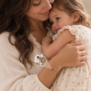 May include: A delicate gold-toned double-strand bracelet featuring a small circular charm with a miniature embedded photograph of a family group. The bracelet has a thin chain design and a single dark circular accent stone next to the photo charm.
