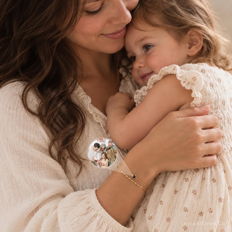 May include: A delicate gold-toned double-strand bracelet featuring a small circular charm with a miniature embedded photograph of a family group. The bracelet has a thin chain design and a single dark circular accent stone next to the photo charm.