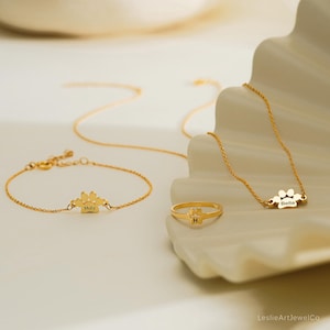 Pode incluir: Um conjunto de joias em tom dourado composto por uma pulseira de corrente delicada com um pingente de pata gravado com o nome Molly, um anel fino com um pequeno la&ccedil;o e a letra H, e um colar com um pingente de pata gravado com o nome Bella.
