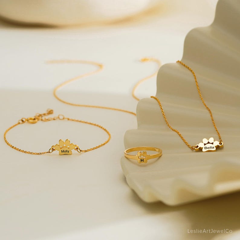 Pode incluir: Um conjunto de joias em tom dourado composto por uma pulseira de corrente delicada com um pingente de pata gravado com o nome Molly, um anel fino com um pequeno la&ccedil;o e a letra H, e um colar com um pingente de pata gravado com o nome Bella.