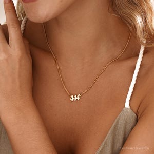May include: A gold-colored necklace with the number "444" pendant. The necklace is worn around the neck. The model is wearing a white spaghetti strap top.