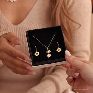 May include: A gold-toned jewelry set consisting of a necklace and two hoop earrings, each featuring a paw print charm. The necklace pendant displays two stacked paw prints, while the earrings each have a single paw print charm. Each charm is engraved with a name.