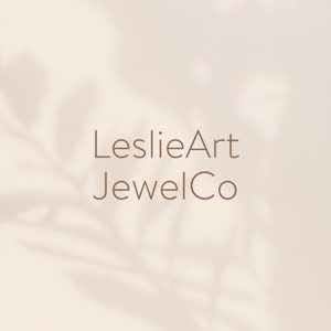 May include: A simple, minimalist logo design with the text "LeslieArt JewelCo" in a light brown color against a white background.