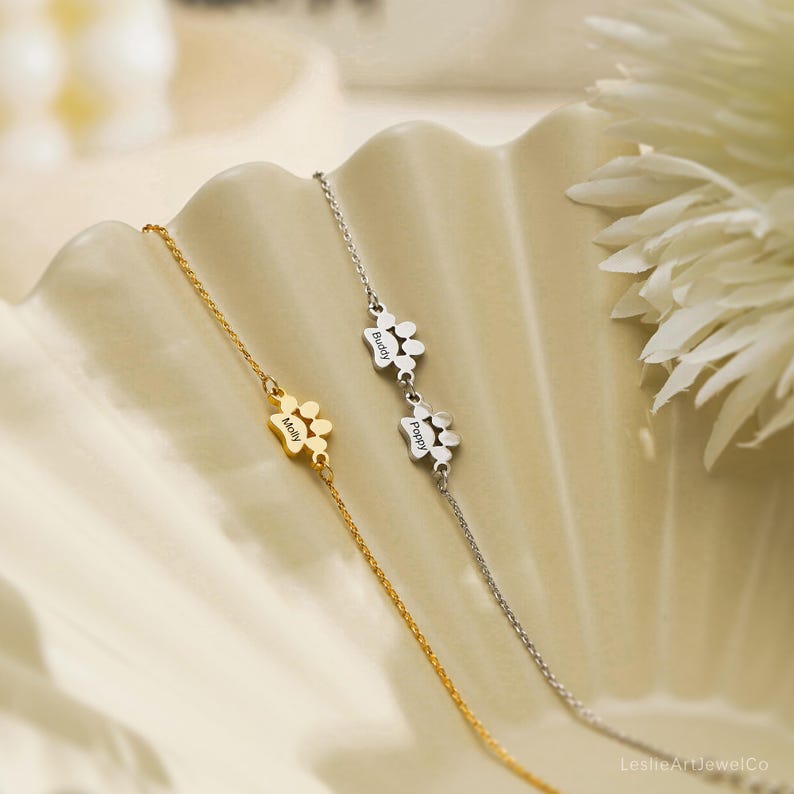 Pode incluir: Duas pulseiras delicadas, uma dourada e outra prateada, cada uma com um pequeno pingente de pata. A pulseira dourada est&aacute; gravada com o nome Molly, enquanto a pulseira prateada apresenta dois pingentes de pata gravados com Buddy e Poppy.