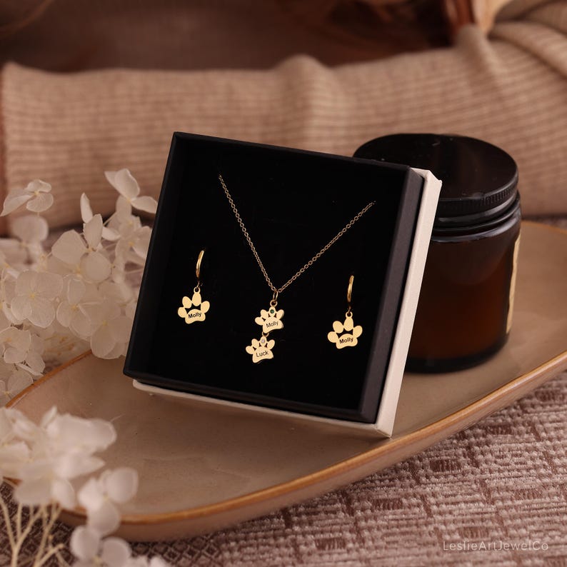 Pode incluir: Um conjunto de joias em tom dourado composto por um colar e dois brincos de argola, cada um adornado com um pingente de pata. O pingente do colar exibe duas patas sobrepostas gravadas com os nomes Molly e Luck, enquanto os brincos apresentam patas individuais gravadas com o nome Molly. O conjunto &eacute; apresentado numa caixa de presente preta.