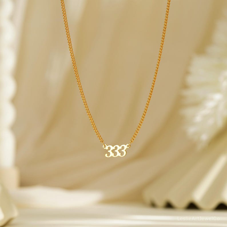 May include: A gold-colored necklace with a delicate chain. The pendant features the number "333" in a stylized font. The necklace is displayed against a soft, neutral background, highlighting the jewelry's design.