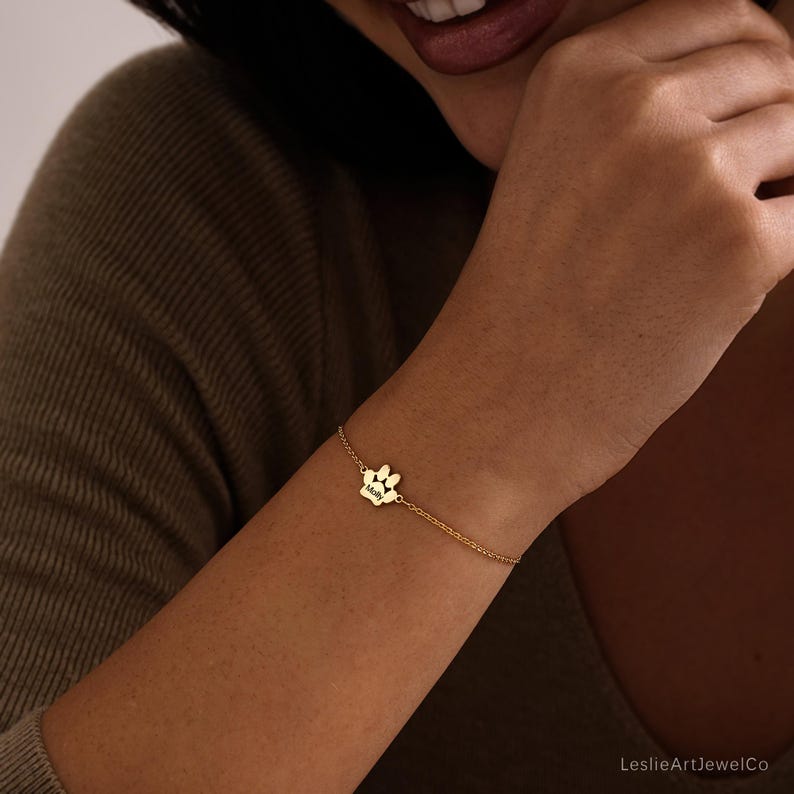Pode incluir: Uma pulseira de corrente em tom dourado com um pingente central em forma de pegada de pata. O pingente de pata tem gravado o nome Molly numa fonte escura e clara. A pulseira consiste numa corrente de metal fina e delicada.