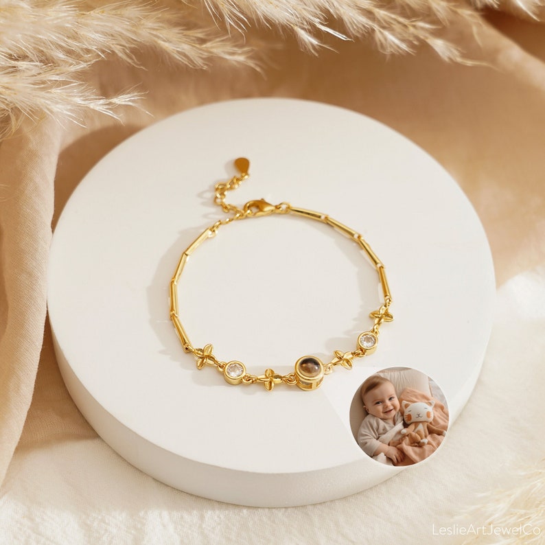 May include: Gold bracelet with a small round locket, surrounded by clear cubic zirconia stones and small flower charms. The locket has a brown background and a small image of a baby.