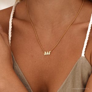 May include: Gold-colored necklace with a delicate chain. The pendant features the number "444" in a simple, elegant font. The necklace is worn against a skin tone background, highlighting the jewelry's design.