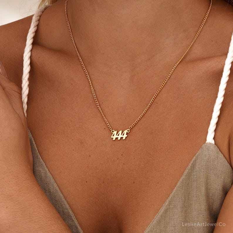 May include: Gold-colored necklace with a delicate chain. The pendant features the number "444" in a simple, elegant font. The necklace is worn against a skin tone background, highlighting the jewelry's design.