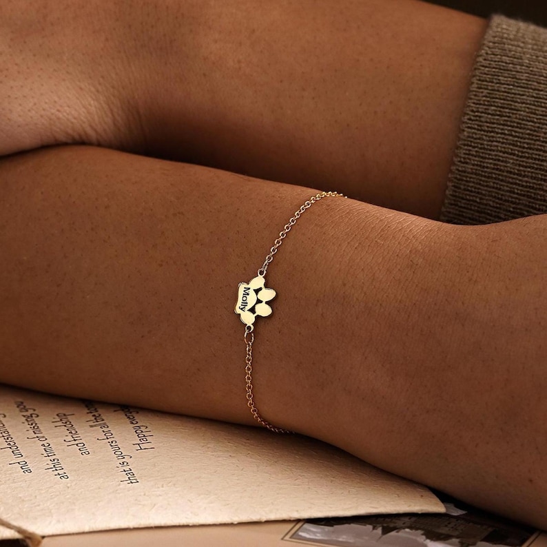 Pode incluir: Uma pulseira de corrente em tom dourado com um pingente central em forma de pegada de animal. O pingente est&aacute; gravado com o nome Molly numa fonte escura e clara. A pulseira consiste numa delicada corrente de elos que prende no pulso.