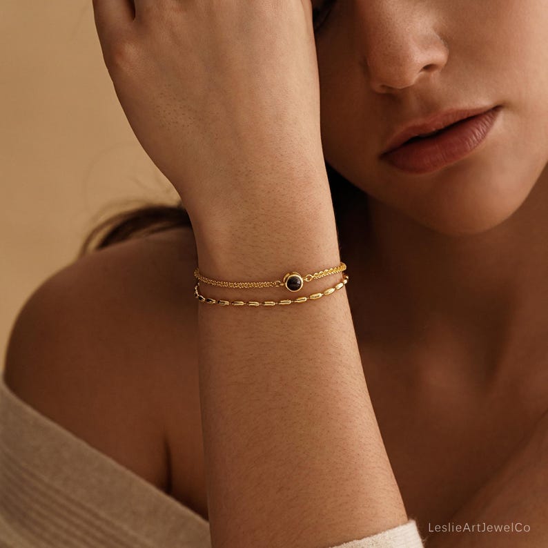 May include: A double-strand gold bracelet featuring a delicate chain with a small, round, dark-colored stone setting and a second chain composed of small, elongated, polished gold beads. The jewelry piece is worn on the wrist.