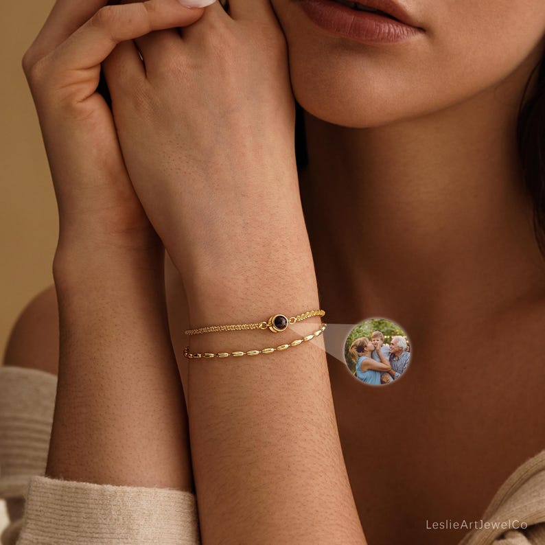 May include: A gold-toned double-strand bracelet featuring a small circular pendant with a dark center. A circular inset displays a color photograph of three people embracing outdoors. The text LeslieArtJewelCo is visible in the bottom right corner.