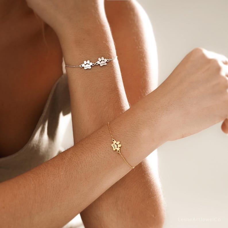 Pode incluir: Uma pulseira de corrente prateada com dois pequenos pingentes de pata com nomes gravados e uma pulseira de corrente dourada com um &uacute;nico pingente de pata pequeno com um nome gravado, usadas nos pulsos.