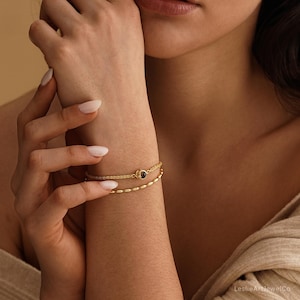 May include: A double-strand gold bracelet featuring a delicate chain with a small circular charm containing a dark center and a second strand composed of small, smooth, oval-shaped gold beads. The jewelry piece is worn on the wrist.