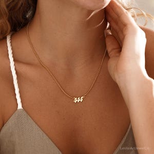 May include: A gold-colored necklace with the number "444" pendant. The necklace has a delicate chain and is worn around the neck. The pendant is in a cursive font. The necklace is a piece of jewelry.