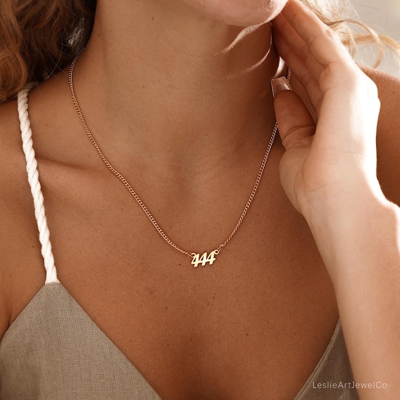 May include: A gold-colored necklace with the number "444" pendant. The necklace has a delicate chain and is worn around the neck. The pendant is in a cursive font. The necklace is a piece of jewelry.