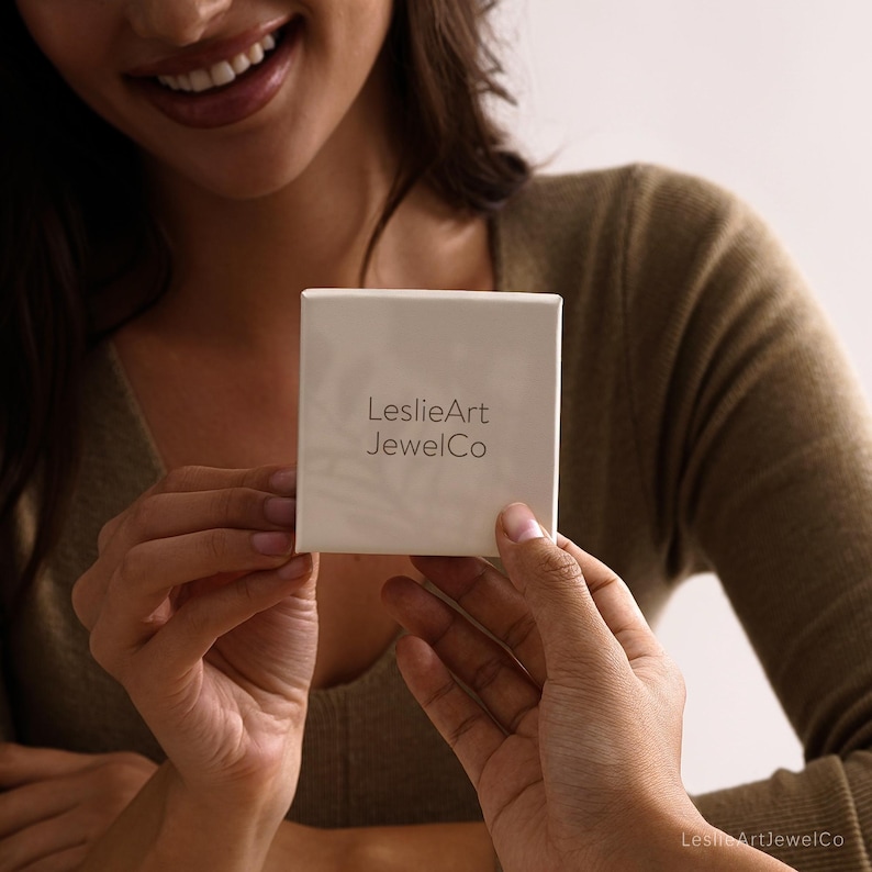 Pode incluir: Uma caixa quadrada de joias em tom branco sujo com o texto LeslieArt JewelCo impresso numa fonte sem serifa cinzento-escura no centro. A caixa tem um acabamento mate suave e um design limpo para guardar pequenos objetos de valor.