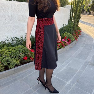 May include: Black skirt with red embroidered detailing on the waist and side panel. The skirt is paired with a black top. The skirt's design features a geometric pattern in red thread. The outfit is worn with black heels.