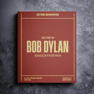 May include: A red book with gold lettering, titled "The Story of Bob Dylan." The cover features the text "Historic Newspapers" and "Daily News." Additional text includes "As Told Through Headlines Since 1963" and "Presented to Simon Branch."