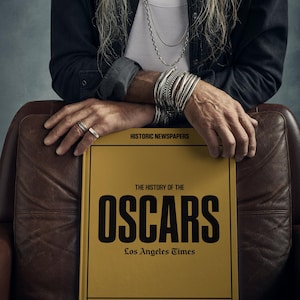 Puede incluir: Un libro amarillo con el t&iacute;tulo "The History of the Oscars" en letras negras. El libro es del Los Angeles Times y presenta titulares de 1920. El libro se presenta a Melissa.