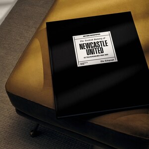 May include: A black hardback book with the title "The Football History of Newcastle United" in white text. The book is on a gold-coloured surface.