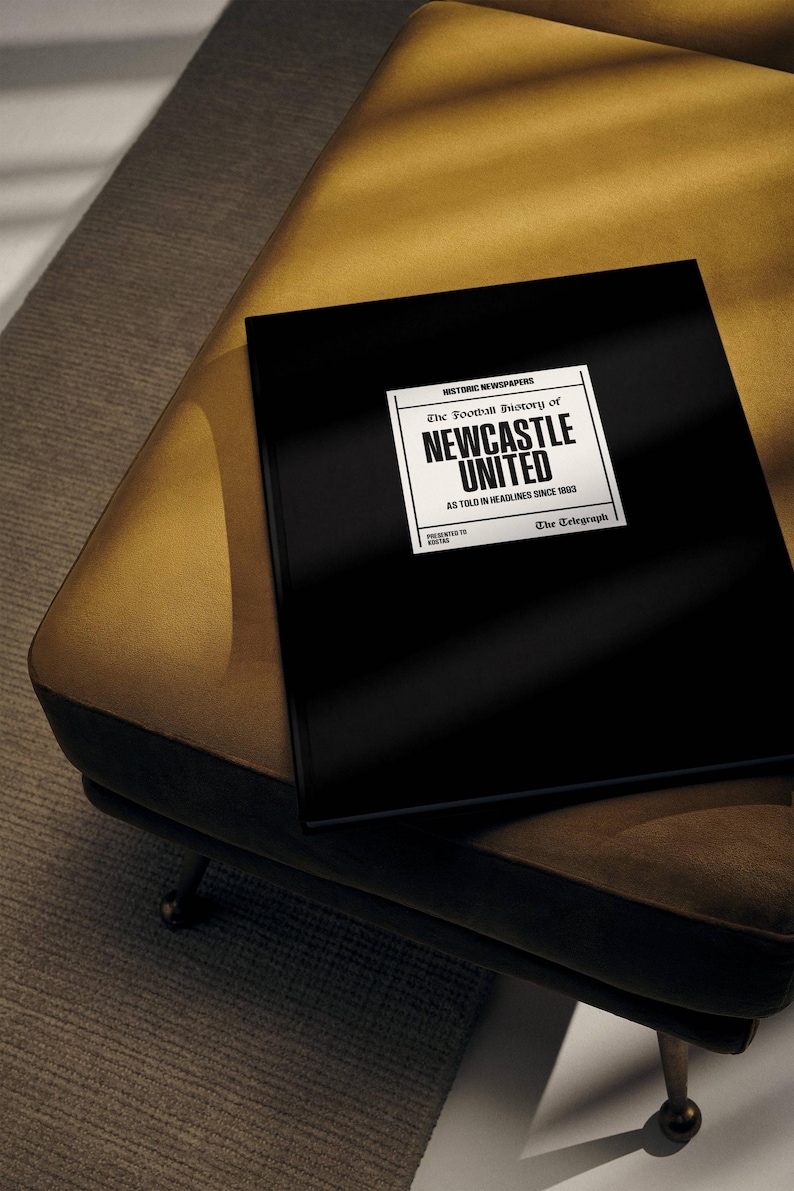 May include: A black hardback book with the title "The Football History of Newcastle United" in white text. The book is on a gold-coloured surface.