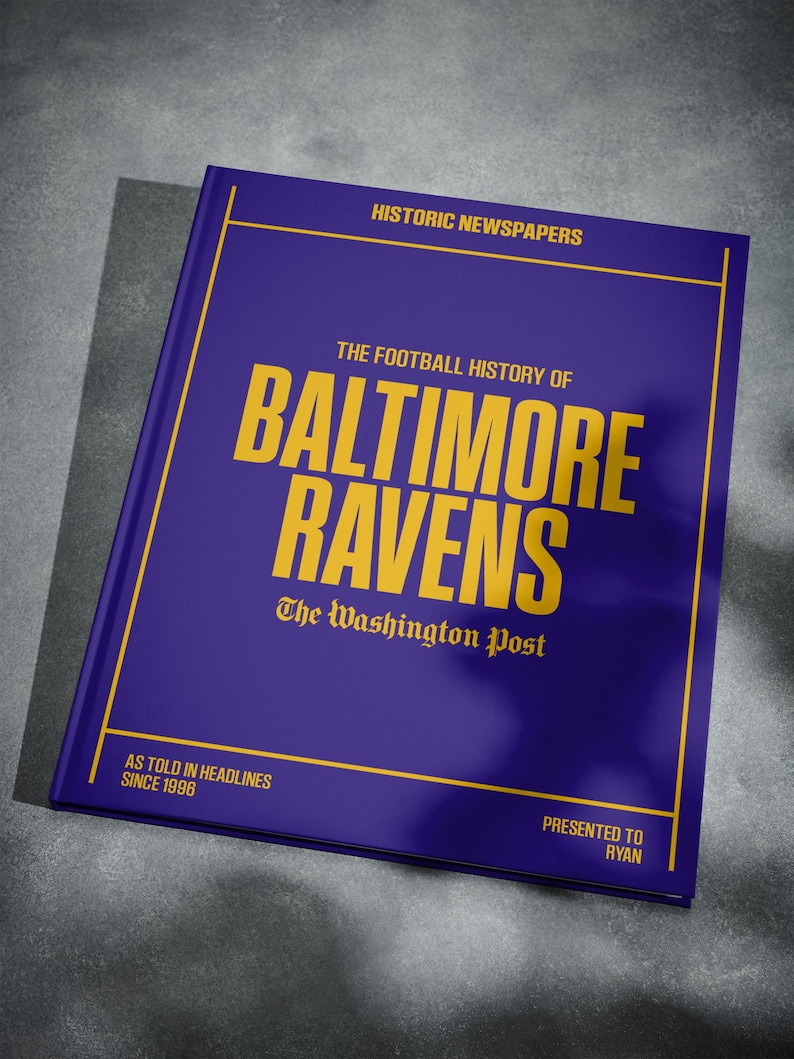 May include: A purple book with the title "The Football History of Baltimore Ravens" in yellow text. The book is presented to Ryan and features the Washington Post logo.