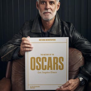 Puede incluir: Un hombre con una chaqueta de cuero marr&oacute;n sostiene un libro blanco titulado "The History of the Oscars" con letras doradas. El libro es publicado por Los Angeles Times y presenta titulares desde 1929.