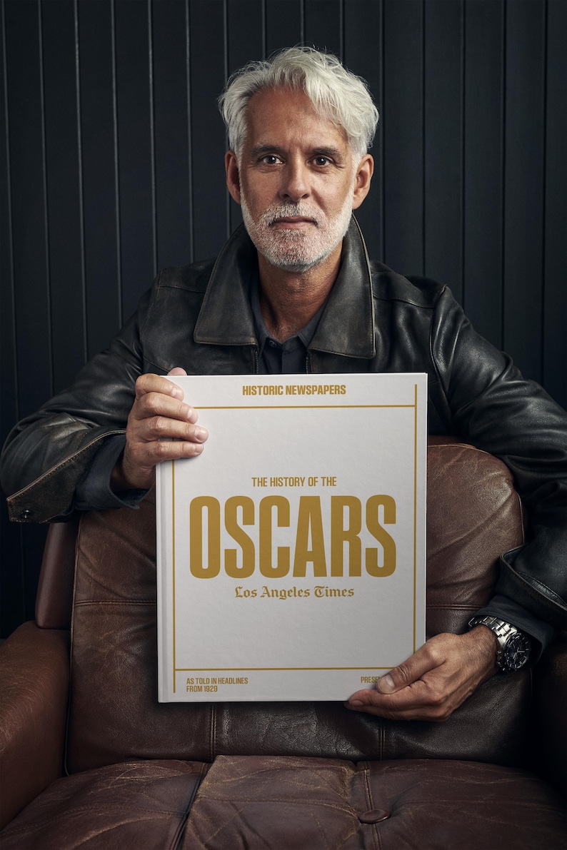 Puede incluir: Un hombre con una chaqueta de cuero marr&oacute;n sostiene un libro blanco titulado "The History of the Oscars" con letras doradas. El libro es publicado por Los Angeles Times y presenta titulares desde 1929.
