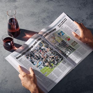 May include: A person reading a newspaper with the headline "Newcastle rampage sends Ashley a timely message". Two glasses of red wine and a decanter of red wine are on a grey surface.