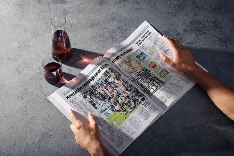 May include: A person reading a newspaper with the headline "Newcastle rampage sends Ashley a timely message". Two glasses of red wine and a decanter of red wine are on a grey surface.