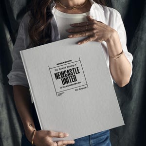 May include: A white book with the title "The Football History of Newcastle United" in black text. The book is held by a person wearing a white shirt and blue jeans.