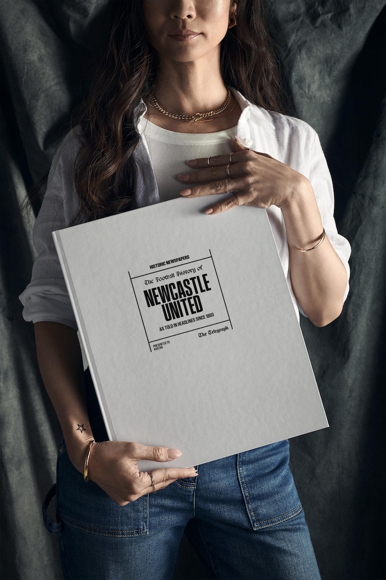 May include: A white book with the title "The Football History of Newcastle United" in black text. The book is held by a person wearing a white shirt and blue jeans.
