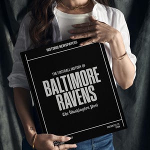 May include: A black book with the title "The Football History of Baltimore Ravens" and the text "The Washington Post" in white lettering. The book is being held by a person wearing a white shirt and blue jeans.