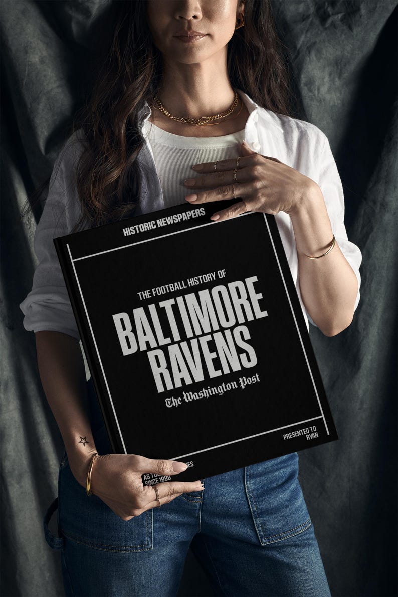 May include: A black book with the title "The Football History of Baltimore Ravens" and the text "The Washington Post" in white lettering. The book is being held by a person wearing a white shirt and blue jeans.