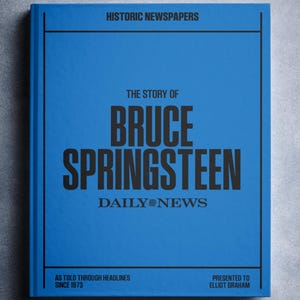 Puede incluir: Portada de libro azul con texto negro: "HISTORIC NEWSPAPERS" y "THE STORY OF BRUCE SPRINGSTEEN" con "DAILY NEWS" debajo. También se ve el texto "AS TOLD THROUGH HEADLINES SINCE 1973" y "PRESENTED TO ELLIOT GRAHAM".