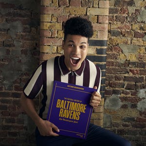 May include: A person wearing a striped shirt holds a purple book with gold lettering that reads "The Fortuitous History of Baltimore Ravens: The Illustrious Past".
