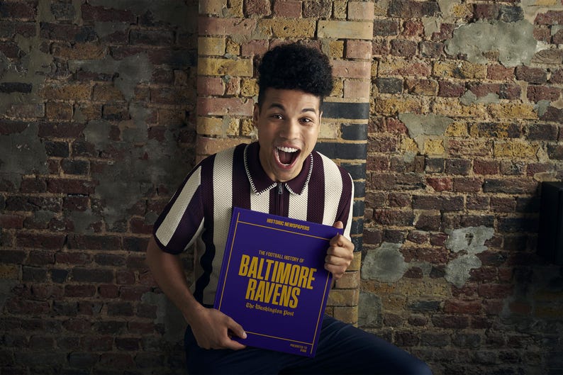 May include: A person wearing a striped shirt holds a purple book with gold lettering that reads "The Fortuitous History of Baltimore Ravens: The Illustrious Past".